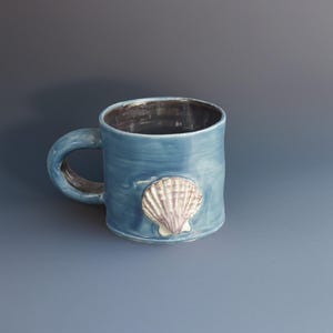 Puede incluir: Una taza de cerámica azul con un diseño de concha. La taza tiene un asa y una superficie texturizada. La concha es blanca y morada. El interior de la taza es oscuro.