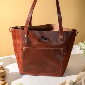 Puede incluir: Un bolso tote de cuero marr&oacute;n con un bolsillo delantero y asas largas. La bolsa tiene el nombre "Olivia" impreso. La bolsa est&aacute; al lado de un par de sandalias beige de tac&oacute;n alto y un ramo de flores blancas.