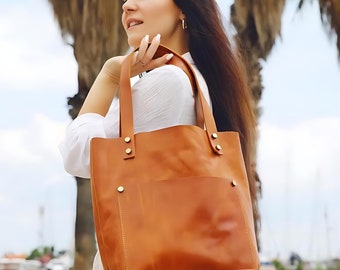 May include: A woman in a white shirt is carrying a brown leather tote bag with a front pocket. The bag has gold accents.