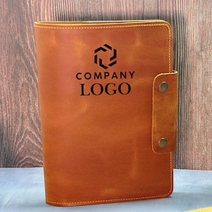 Peut inclure: Un journal en cuir de couleur fauve avec une fermeture à boucle. La couverture présente un logo d'entreprise noir et les mots "COMPANY LOGO". Le journal est fermé par une lanière en cuir et deux rivets couleur laiton.
