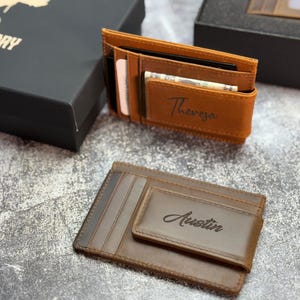 May include: Two leather wallets, one tan and one brown, with personalized names. The tan wallet has "Theresa" engraved, and the brown wallet has "Austin" engraved. Both wallets are open, revealing card slots and cash.