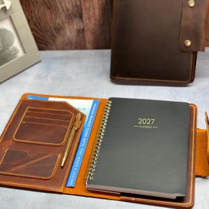 May include: A brown leather planner cover featuring an open interior with card slots, a gold pen, and a spiral-bound 2027 planner. A closed brown leather cover with two brass snap buttons sits in the background next to a gray picture frame.