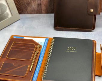 May include: A brown leather planner cover featuring an open interior with card slots, a gold pen, and a spiral-bound 2027 planner. A closed brown leather cover with two brass snap buttons sits in the background next to a gray picture frame.
