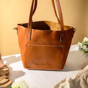 Puede incluir: Un bolso tote de cuero marr&oacute;n con un bolsillo frontal y asas largas. La palabra "amanda" est&aacute; escrita en cursiva en el bolsillo frontal. Un par de sandalias beige de tac&oacute;n alto y flores est&aacute;n al lado del bolso.