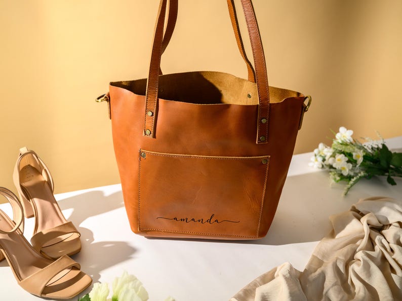 Puede incluir: Un bolso tote de cuero marr&oacute;n con un bolsillo frontal y asas largas. La palabra "amanda" est&aacute; escrita en cursiva en el bolsillo frontal. Un par de sandalias beige de tac&oacute;n alto y flores est&aacute;n al lado del bolso.