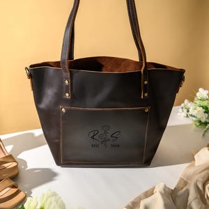Puede incluir: Un bolso tote de cuero marr&oacute;n oscuro con un bolsillo delantero y asas. El bolso tiene un dise&ntilde;o floral y el texto "Rose Shaw" impreso en la parte delantera. Un par de sandalias beige de tac&oacute;n alto y un ramo de flores blancas est&aacute;n junto al bolso.