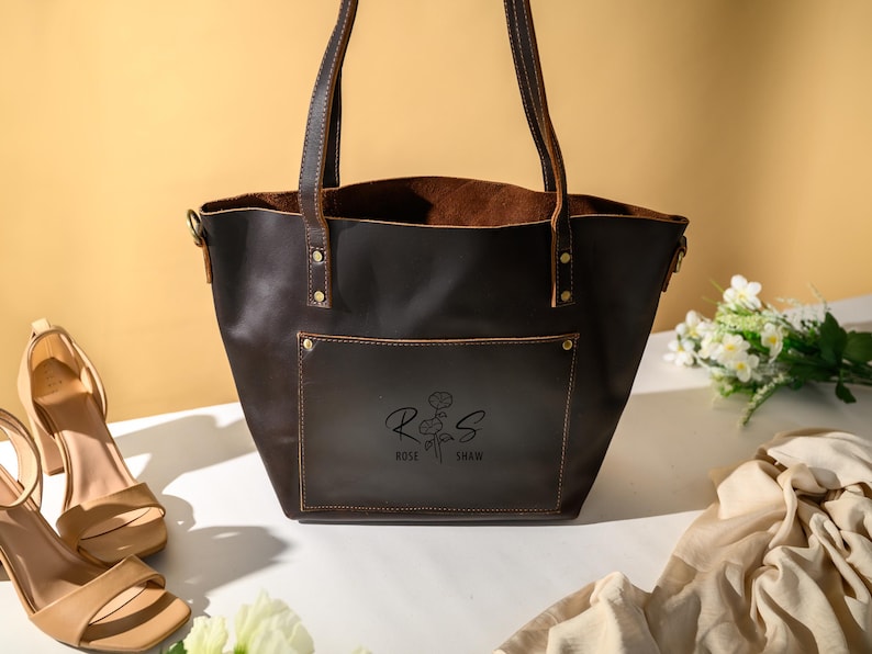 Puede incluir: Un bolso tote de cuero marr&oacute;n oscuro con un bolsillo delantero y asas. El bolso tiene un dise&ntilde;o floral y el texto "Rose Shaw" impreso en la parte delantera. Un par de sandalias beige de tac&oacute;n alto y un ramo de flores blancas est&aacute;n junto al bolso.