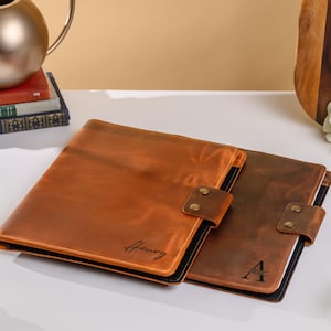 May include: Two leather folios in shades of brown, one lighter and one darker, are displayed. Each folio has a flap closure with a brass-colored snap. The lighter folio is inscribed with the name "Henry", while the darker one has the initial "A".