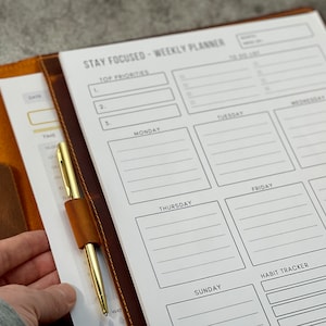 May include: A weekly planner with the text "STAY FOCUSED - WEEKLY PLANNER" is open to a blank page. The planner is in a brown leather cover with a gold pen secured by a leather loop. The planner has sections for each day of the week, top priorities, and a habit tracker.