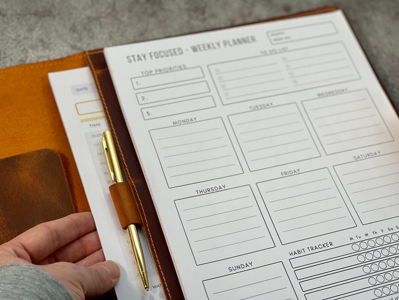May include: A weekly planner with the text "STAY FOCUSED - WEEKLY PLANNER" is open to a blank page. The planner is in a brown leather cover with a gold pen secured by a leather loop. The planner has sections for each day of the week, top priorities, and a habit tracker.