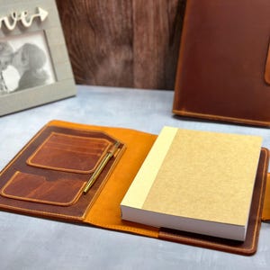 Peut inclure: Une couverture de carnet en cuir marron pr&eacute;sentant un int&eacute;rieur ouvert avec deux fentes pour cartes, un porte-stylo contenant un stylo dor&eacute; et un carnet &eacute;pais &agrave; couverture beige. Une couverture de carnet ferm&eacute;e assortie avec bouton-pression.