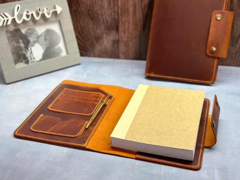 Peut inclure: Une couverture de carnet en cuir marron pr&eacute;sentant un int&eacute;rieur ouvert avec deux fentes pour cartes, un porte-stylo contenant un stylo dor&eacute; et un carnet &eacute;pais &agrave; couverture beige. Une couverture de carnet ferm&eacute;e assortie avec bouton-pression.
