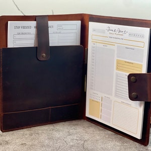 May include: Open leather planner with a dark brown cover and a light brown interior. The planner includes a daily planner pad with sections for activity, focus, and notes. A pocket holds additional papers. The text "Stay Focused - Weekly Planner" is visible.