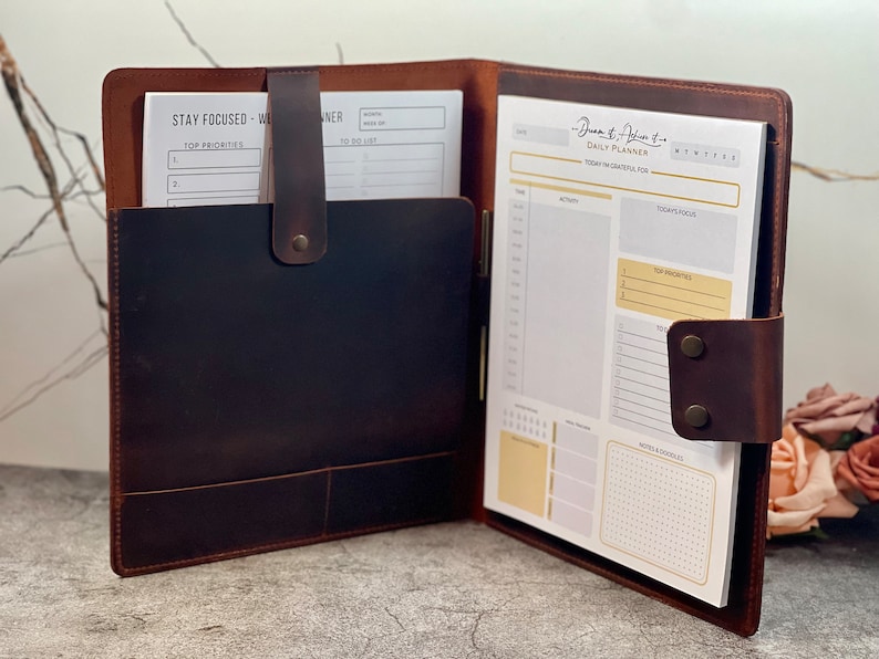 May include: Open leather planner with a dark brown cover and a light brown interior. The planner includes a daily planner pad with sections for activity, focus, and notes. A pocket holds additional papers. The text "Stay Focused - Weekly Planner" is visible.