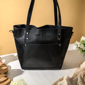 Puede incluir: Un bolso tote de cuero negro con un bolsillo frontal y correas para los hombros. La bolsa tiene el nombre "KATHY Morgan" impreso en la parte delantera. Un par de sandalias de tac&oacute;n alto color beige y un ramo de flores blancas est&aacute;n junto a la bolsa.