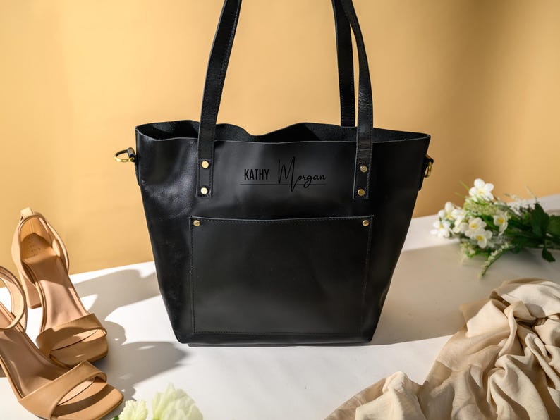 Puede incluir: Un bolso tote de cuero negro con un bolsillo frontal y correas para los hombros. La bolsa tiene el nombre "KATHY Morgan" impreso en la parte delantera. Un par de sandalias de tac&oacute;n alto color beige y un ramo de flores blancas est&aacute;n junto a la bolsa.