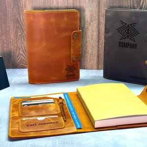 Peut inclure: Deux prot&egrave;ge-carnets en cuir, l'un beige et l'autre marron fonc&eacute;, dot&eacute;s d'une fermeture &agrave; pression et d'un logo g&eacute;om&eacute;trique. Un prot&egrave;ge-carnet ouvert pr&eacute;sente un bloc-notes jaune, un stylo et des fentes pour cartes avec le nom Carl Johnson.