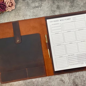 May include: A brown leather weekly planner with a pen holder and a gold pen. The planner is open, revealing a white paper with a "Stay Focused" layout for priorities, to-do lists, and a habit tracker. The planner is closed with a snap closure.