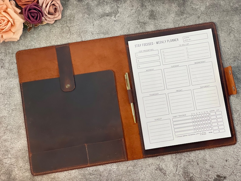 May include: A brown leather weekly planner with a pen holder and a gold pen. The planner is open, revealing a white paper with a "Stay Focused" layout for priorities, to-do lists, and a habit tracker. The planner is closed with a snap closure.