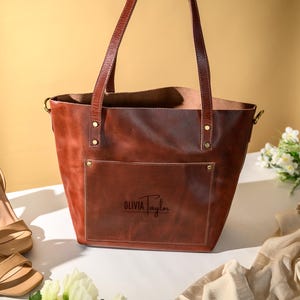 Puede incluir: Un bolso tote de cuero marr&oacute;n con asas largas y un bolsillo delantero. El bolso tiene forma rectangular y est&aacute; personalizado con el nombre "Olivia Taylor". Sandalias de tac&oacute;n beige y flores est&aacute;n al lado del bolso.