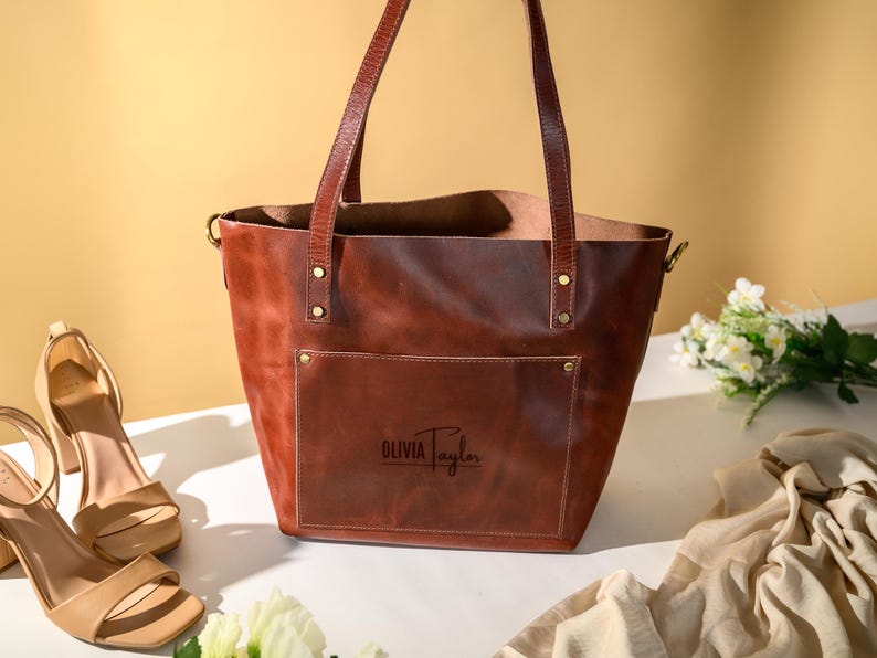 Puede incluir: Un bolso tote de cuero marr&oacute;n con asas largas y un bolsillo delantero. El bolso tiene forma rectangular y est&aacute; personalizado con el nombre "Olivia Taylor". Sandalias de tac&oacute;n beige y flores est&aacute;n al lado del bolso.