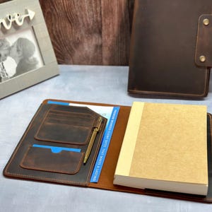 May include: A brown leather notebook cover with a pen holder, multiple pockets, and a flap closure. The cover is open to reveal a blank notebook.