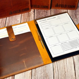 May include: A brown leather portfolio with a yellow interior and a weekly planner insert. The planner has a section for daily tasks, a habit tracker, and a section for top priorities. The portfolio has the text "FIELD & FARM" embossed on the front.