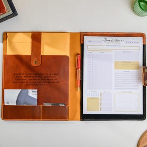 May include: A brown leather planner, open to reveal a daily planner pad and a red pen. The planner has a yellow interior and a quote: "SUCCESS IS NO ACCIDENT. IT IS HARD WORK, PERSEVERANCE, LEARNING..." The pad is for daily planning.
