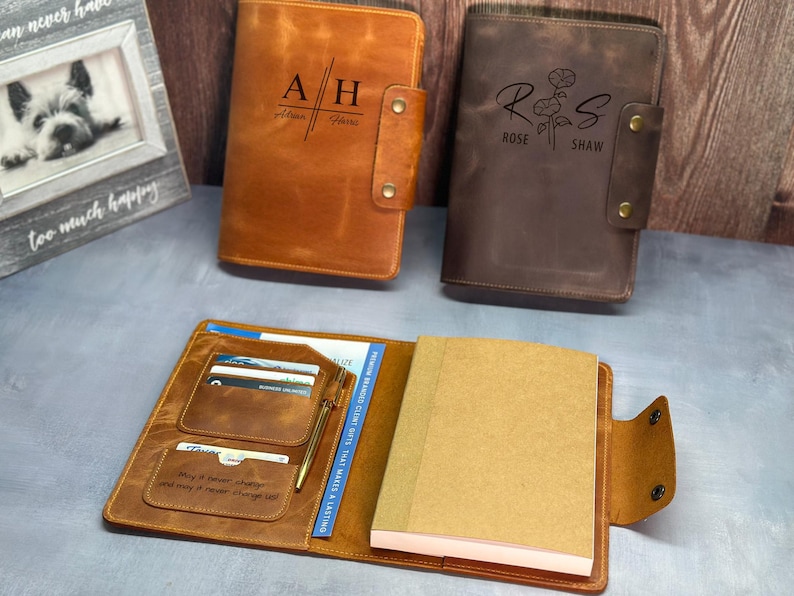 May include: Two leather-bound journals with personalized engravings. The brown journal has the initials "AH" and the name "Alan Harris". The dark brown journal has the initials "RS" and the name "Rose Shaw" with a floral design. Both journals have a pen holder and card slots.