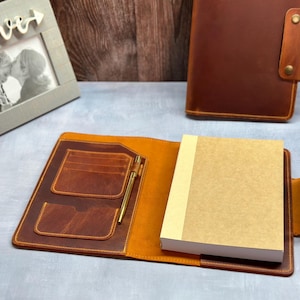 May include: A brown leather notebook with a pen holder and a pocket for cards. The notebook is open and has a blank page inside.
