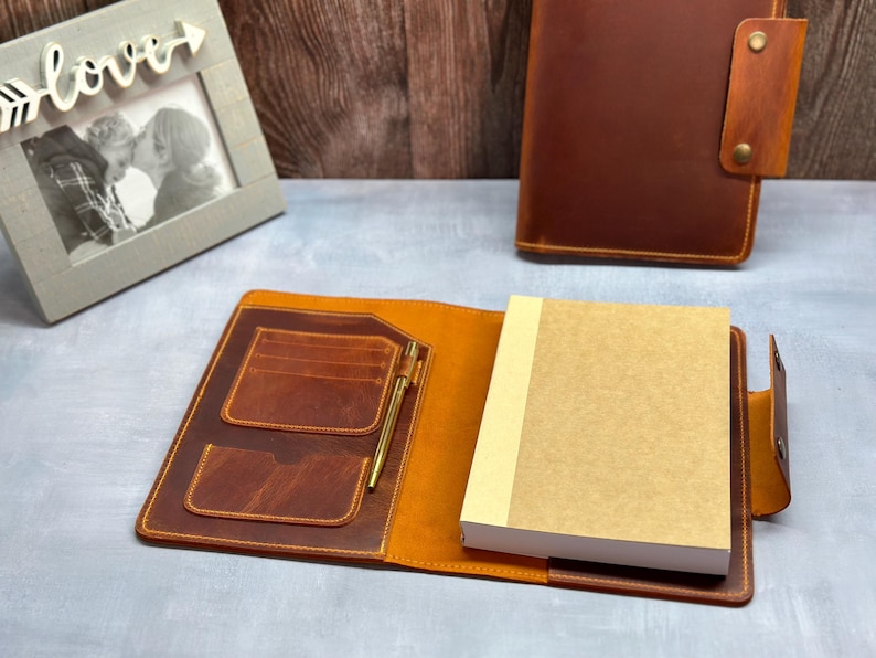 May include: A brown leather notebook with a pen holder and a pocket for cards. The notebook is open and has a blank page inside.
