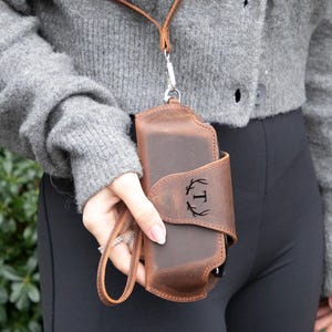May include: A brown leather phone case with a strap and a monogrammed initial "T" in a wreath design.