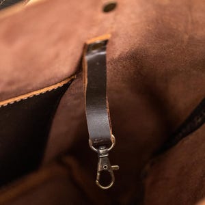 Puede incluir: Primer plano del interior de un bolso tote de cuero marr&oacute;n. El bolso presenta una correa de cuero marr&oacute;n oscuro con un cierre de color plateado. El interior es de un tono marr&oacute;n m&aacute;s claro, con costuras visibles y un cierre de bot&oacute;n.