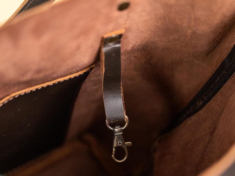 Puede incluir: Primer plano del interior de un bolso tote de cuero marr&oacute;n. El bolso presenta una correa de cuero marr&oacute;n oscuro con un cierre de color plateado. El interior es de un tono marr&oacute;n m&aacute;s claro, con costuras visibles y un cierre de bot&oacute;n.