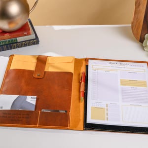 May include: Open leather portfolio with a notepad, pen, and card holder. The portfolio is brown with a yellow interior and a black border around the notepad. The notepad has a daily planner layout. A quote is embossed on the portfolio.