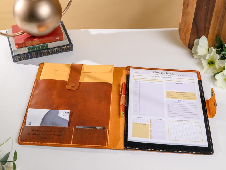 May include: Open leather portfolio with a notepad, pen, and card holder. The portfolio is brown with a yellow interior and a black border around the notepad. The notepad has a daily planner layout. A quote is embossed on the portfolio.