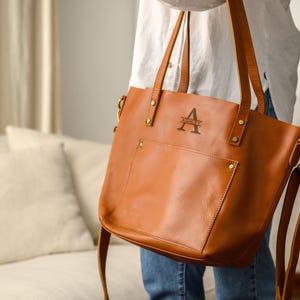 Puede incluir: Un bolso tote de cuero marr&oacute;n con una correa para el hombro y asas superiores. El bolso tiene un bolsillo frontal y un monograma con la letra "A" y el nombre "Ashley". El bolso lo sostiene una persona que lleva una camisa blanca y vaqueros.