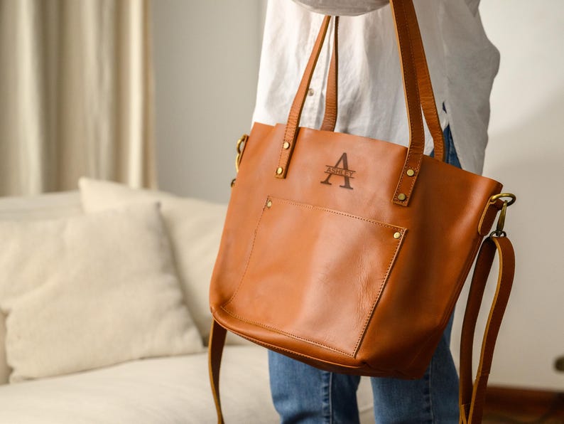 Puede incluir: Un bolso tote de cuero marr&oacute;n con una correa para el hombro y asas superiores. El bolso tiene un bolsillo frontal y un monograma con la letra "A" y el nombre "Ashley". El bolso lo sostiene una persona que lleva una camisa blanca y vaqueros.