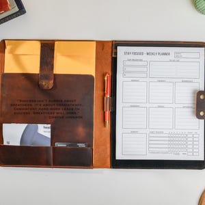 May include: Open leather planner with a weekly layout and a pen. The planner is brown with a quote from Dwayne Johnson. The left side has pockets and a yellow envelope. The right side has a weekly planner page.