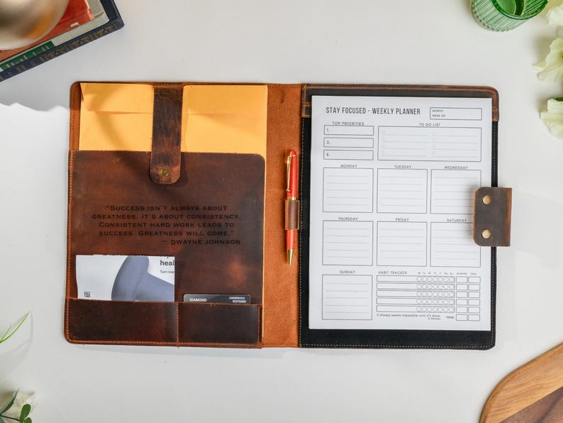 May include: Open leather planner with a weekly layout and a pen. The planner is brown with a quote from Dwayne Johnson. The left side has pockets and a yellow envelope. The right side has a weekly planner page.