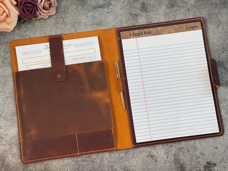 May include: Open leather portfolio in shades of brown, featuring a notepad, pen, and a pocket for documents. The notepad has blue lines and the words "Legal Pad" printed at the top. A gold pen is inserted in a leather loop.