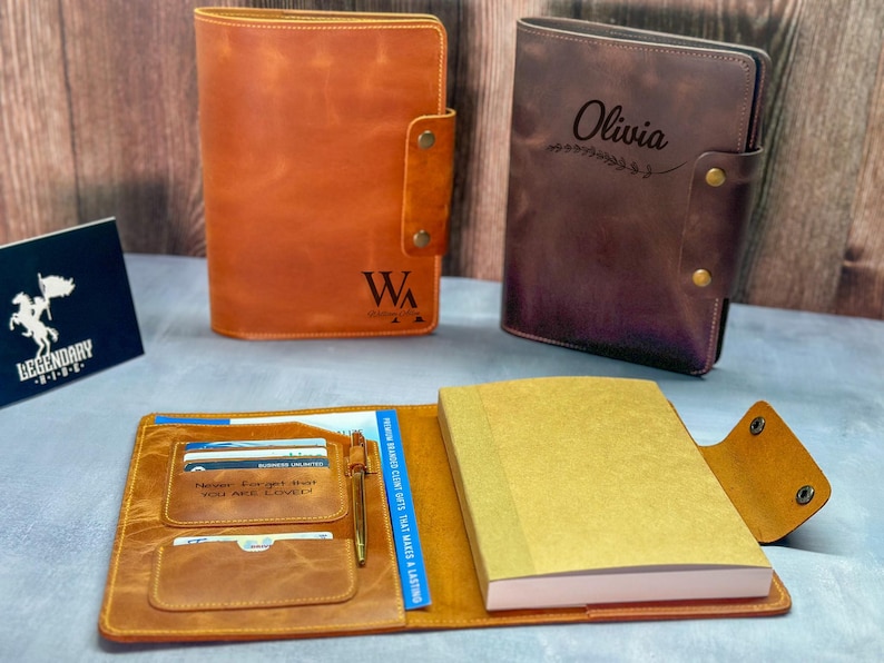 Peut inclure: Deux couvertures de journal en cuir, l'une fauve et l'autre brun fonc&eacute;, avec les initiales WA et le pr&eacute;nom Olivia. Une couverture fauve ouverte pr&eacute;sente un carnet, des fentes pour cartes, un stylo et le texte grav&eacute; Never Forget That You Are Loved.