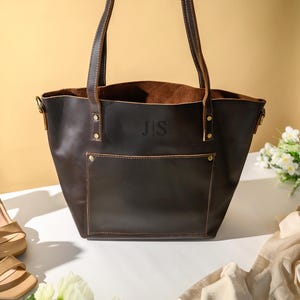 Puede incluir: Un bolso tote de cuero marr&oacute;n oscuro con un bolsillo frontal y dos asas. La bolsa tiene las iniciales "J S" grabadas en relieve en la parte delantera. Sandalias de tac&oacute;n alto beige y una bufanda color crema est&aacute;n junto a la bolsa. Tambi&eacute;n hay flores blancas en la imagen.