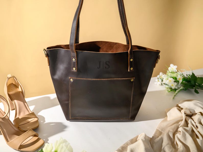Puede incluir: Un bolso tote de cuero marr&oacute;n oscuro con un bolsillo frontal y dos asas. La bolsa tiene las iniciales "J S" grabadas en relieve en la parte delantera. Sandalias de tac&oacute;n alto beige y una bufanda color crema est&aacute;n junto a la bolsa. Tambi&eacute;n hay flores blancas en la imagen.