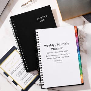 May include: A black spiral-bound 2027 planner with white text on the cover and an open white weekly/monthly planner with colorful monthly tabs. A rectangular invitation card with gold borders and black text is positioned underneath the planners.