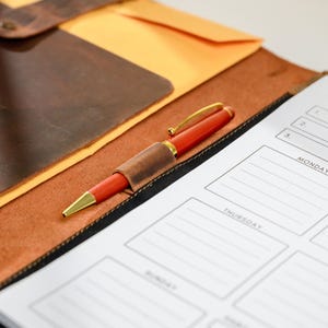 May include: A red pen with gold accents rests on a leather notebook with a matching pen holder. A planner with the words "Stay Focused" and daily sections is visible. The notebook and planner are on a brown leather surface.