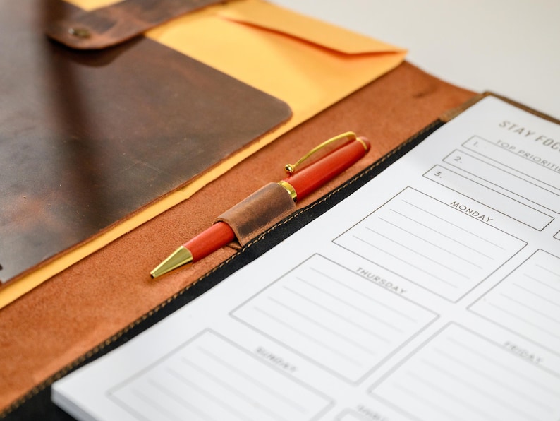 May include: A red pen with gold accents rests on a leather notebook with a matching pen holder. A planner with the words "Stay Focused" and daily sections is visible. The notebook and planner are on a brown leather surface.