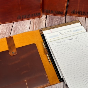 May include: Leather planner with a brown cover and a light brown interior. The planner includes a notepad, pen, and a weekly planner. The text "FIELD & FARM" is embossed on the cover. The notepad has the text "Legal Pad" and "STAY FOCUSED - WEEKLY PLANNER".