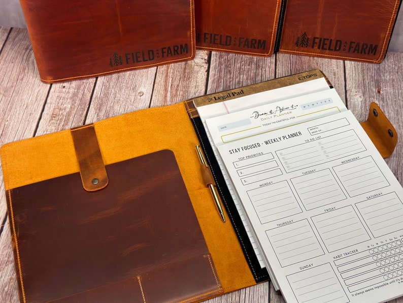 May include: Leather planner with a brown cover and a light brown interior. The planner includes a notepad, pen, and a weekly planner. The text "FIELD & FARM" is embossed on the cover. The notepad has the text "Legal Pad" and "STAY FOCUSED - WEEKLY PLANNER".