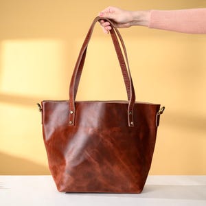 Puede incluir: Un bolso tote de cuero marr&oacute;n con asas largas. El bolso es de un color marr&oacute;n rojizo intenso y parece estar hecho de cuero de alta calidad. Las asas est&aacute;n sujetas con herrajes met&aacute;licos. El bolso es sostenido por una persona.